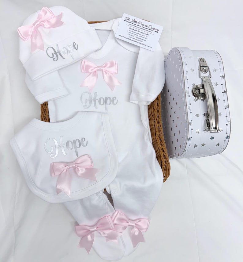 May include: A white baby set including a long-sleeve footed sleepsuit, a bib, and a hat, all featuring the name Hope in silver script and light pink satin bows. The set is presented in a wicker basket with a white suitcase featuring silver stars.