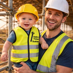 May include: A bright yellow high-visibility safety vest for a toddler featuring black trim and the text Daddy's Trainee Scaffolder with a wrench icon. The vest includes horizontal reflective silver stripes and is worn over a blue shirt.