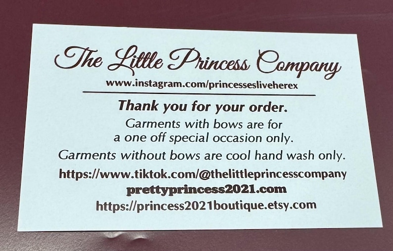 May include: A rectangular white card with dark brown text that reads The Little Princess Company, followed by social media links, a thank you message, and care instructions for garments with and without bows, including website addresses.