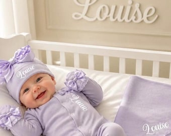 May include: A light purple baby outfit consisting of a long-sleeve onesie with a front button closure, a matching hat with a large bow, and a blanket. Each item features the name Louise embroidered in white script, with bows on the cuffs.