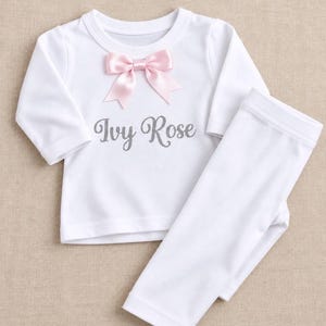May include: A white long-sleeve shirt and matching trousers set. The shirt features a light pink satin bow at the neckline and the name Ivy Rose printed in a grey script font across the centre of the chest area.