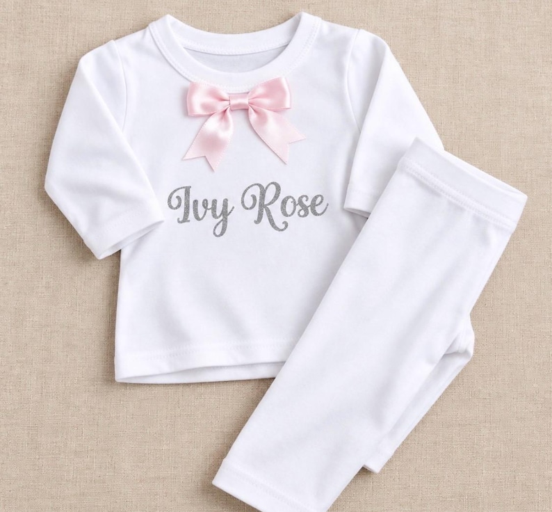 May include: A white long-sleeve shirt and matching trousers set. The shirt features a light pink satin bow at the neckline and the name Ivy Rose printed in a grey script font across the centre of the chest area.