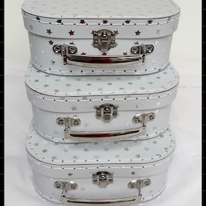 May include: A stack of three white suitcases with silver star patterns, metal latches, and silver handles. The text reads: Exclusively designed by me! Why not add a gift bag Which comes as a keepsake memory suitcase.