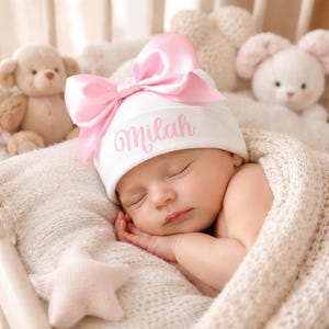 May include: A white newborn beanie featuring a large, shiny pink satin bow attached to the front. The hat is embroidered with the name Milah in a pink cursive script font. The beanie is made of a soft fabric and is designed to fit a small head.