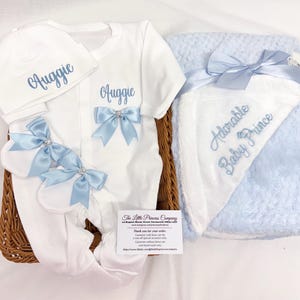 Puede incluir: Un conjunto de regalo para bebé blanco con gorro, body y manoplas, todos bordados con el nombre "Auggie" en azul. El body y las manoplas tienen lazos azules con detalles de pedrería. También se incluye una manta azul y blanca con el bordado "Adorable Baby Prince".