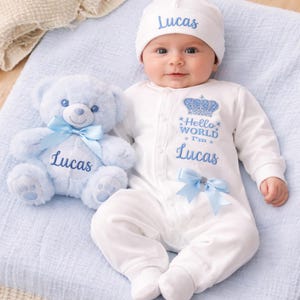 May include: A white baby onesie with long sleeves and a matching white beanie, both embroidered with the name Lucas in blue. The onesie features a blue crown graphic, the text Hello WORLD I'm Lucas, and a light blue ribbon bow at the waist.