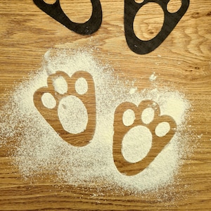 May include: Two black paw print stencils on a wooden surface. The stencils are placed on top of a white powder, creating an imprint of the paw prints.