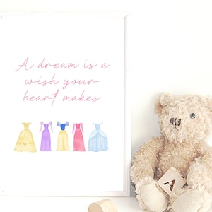 May include: A framed print featuring the quote "A dream is a wish your heart makes" in pink script. Below are watercolour illustrations of five princess dresses in yellow, purple, yellow, pink, and blue. A teddy bear sits to the right.