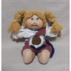 Puede incluir: Muñeca Cabbage Patch Kid vintage con coletas rubias de hilo y ojos azules. La muñeca lleva una camisa blanca, una falda a cuadros rojos y verdes y una bolsa marrón. Los brazos y las piernas de la muñeca son de color beige claro.