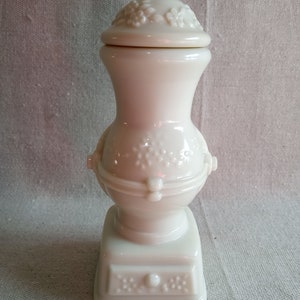 Puede incluir: Botella de porcelana blanca con un diseño floral en la tapa y un diseño de copo de nieve en el cuerpo. La botella tiene una base redonda y una parte superior cuadrada.