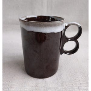 May include: A brown ceramic mug with a distinctive handle. The mug has a dark brown exterior and a white rim. The handle has two circular openings. The mug is designed for hot drinks.