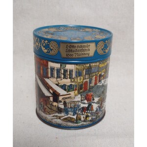 May include: A blue cylindrical tin featuring a detailed illustration of a European town scene. The tin has the text "E-Otto Schmidt Lebkuchenfabrik 8500 Nürnberg" in gold lettering. The lid is decorated with a floral pattern.