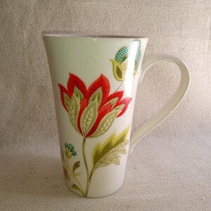 May include: White ceramic mug with a tall, slender shape and a large handle. The mug features a floral design with red, green, and gold accents.