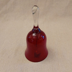 May include: A red glass bell with a clear glass handle. The bell has a shiny, reflective surface.
