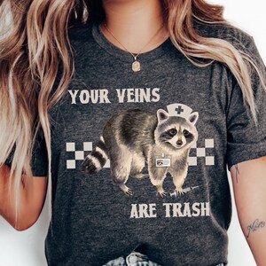 May include: A dark grey heather t-shirt with a graphic of a raccoon wearing a nurse's hat and holding a syringe. The text "Your Veins Are Trash" is printed on the shirt.