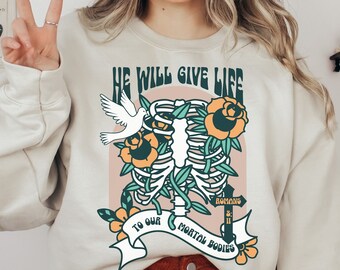May include: A beige sweatshirt with a graphic print of a ribcage with flowers and a dove. The text on the graphic reads "He will give life to our mortal bodies, Romans 8:11".