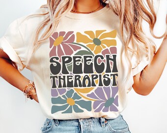May include: A white t-shirt with a retro floral design and the text "SPEECH THERAPIST" in a groovy font.