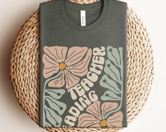 May include: A heather military green t-shirt with a floral design and the text "Reading Teacher" in a retro font.