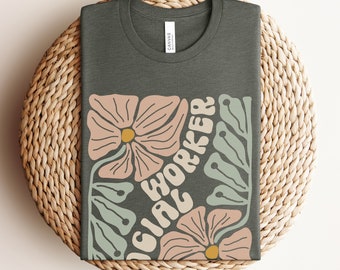 May include: A heather military green t-shirt with a floral design and the text "Social Worker" in a retro font.