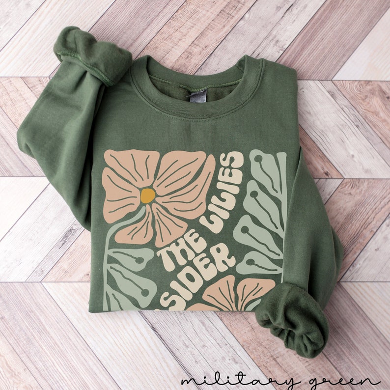 K&ouml;nnte beinhalten: Ein gr&uuml;ner Pullover mit einem Blumendesign und dem Text "The Sider Lilies" in einer Retro-Schrift.