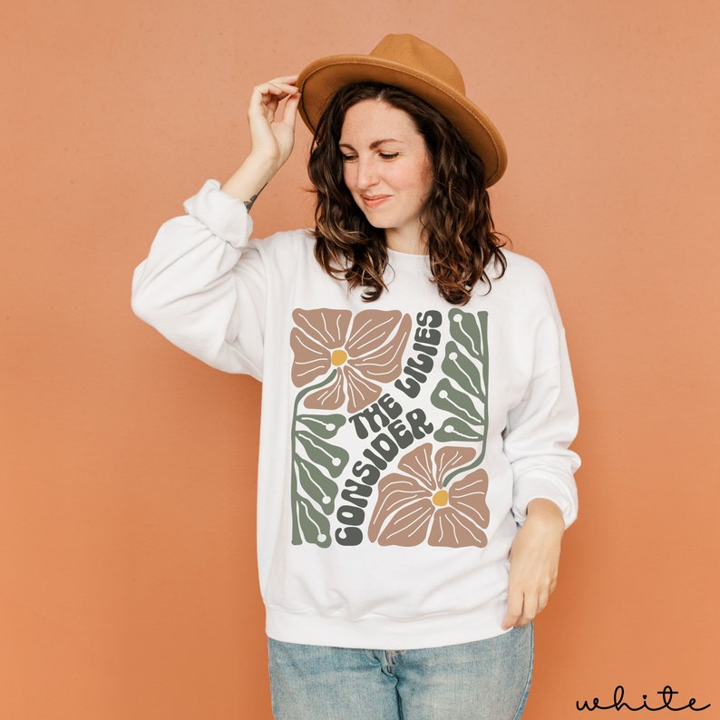 K&ouml;nnte beinhalten: Wei&szlig;er Pullover mit einem Blumendesign und dem Text "Consider the lilies" in einer Retro-Schrift.