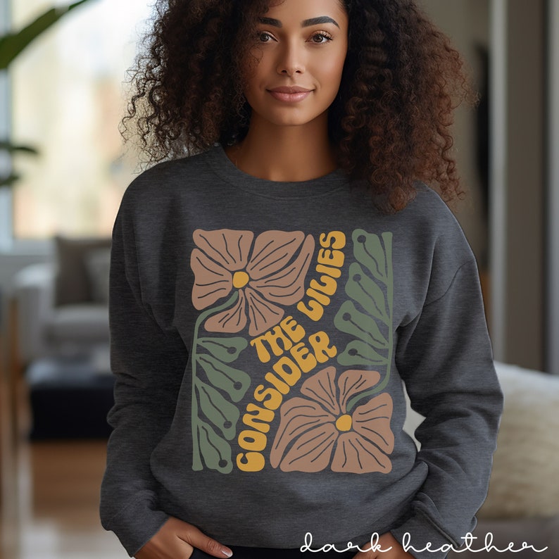 K&ouml;nnte beinhalten: Dunkelgrauer Sweatshirt mit einem Blumendesign und dem Text "Consider the Lilies" in einer Retro-Schrift.