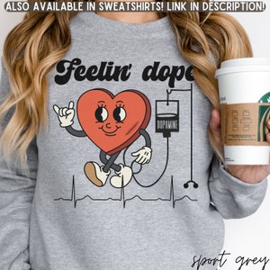 May include: A gray sweatshirt with a red heart graphic. The heart has a smiling face, arms, and legs. The heart is connected to an IV bag labeled "Dopamine". The text "Feelin' dope" is above the heart.