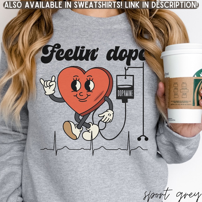 May include: A gray sweatshirt with a red heart graphic. The heart has a smiling face, arms, and legs. The heart is connected to an IV bag labeled "Dopamine". The text "Feelin' dope" is above the heart.