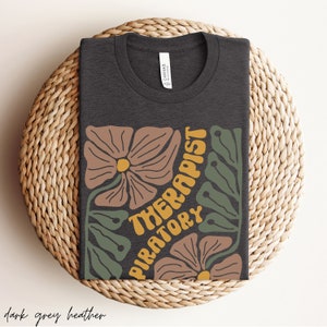 May include: Dark grey heather t-shirt with a floral design and the text "Piratory Therapist" in yellow.