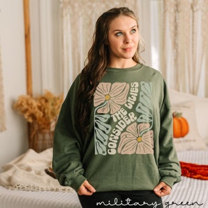 K&ouml;nnte beinhalten: Ein gr&uuml;nes Sweatshirt mit einem Blumendesign und dem Text "Consider the lilies" in einer Retro-Schrift.