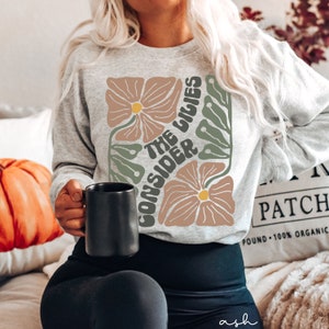 K&ouml;nnte beinhalten: Ein graues Sweatshirt mit einem floralen Design und dem Text "Consider the lilies" in einer Retro-Schrift.