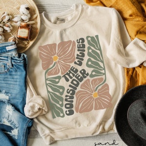 K&ouml;nnte beinhalten: Ein beiger Pullover mit einem Blumendesign und dem Text "Consider the lilies" in einer Retro-Schrift.