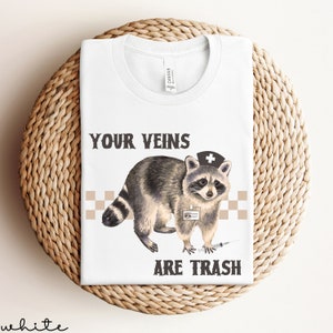 May include: White t-shirt with a raccoon wearing a nurse's hat and holding a syringe. The text "YOUR VEINS ARE TRASH" is printed on the shirt.