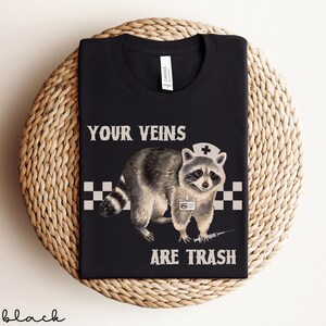 May include: Black t-shirt with a graphic of a raccoon wearing a nurse's hat and holding a syringe. The text "YOUR VEINS ARE TRASH" is printed on the shirt.