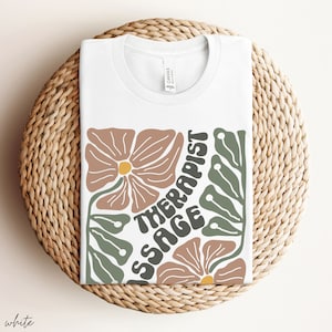 May include: White t-shirt with a floral design and the text "Massage Therapist" in a retro font.