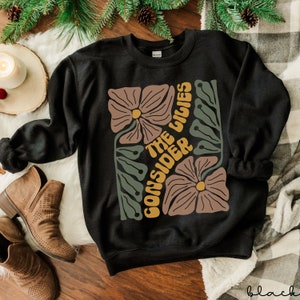 K&ouml;nnte beinhalten: Schwarzer Pullover mit einem Blumendesign und dem Text "Consider the lilies" in Gelb.