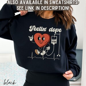 May include: Black sweatshirt with a graphic of a red heart with a smiling face and a bandage, connected to an IV bag labeled "Dopamine". The text "Feelin' dope" is above the heart graphic.