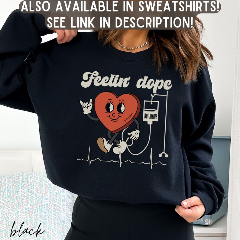 May include: Black sweatshirt with a graphic of a red heart with a smiling face and a bandage, connected to an IV bag labeled "Dopamine". The text "Feelin' dope" is above the heart graphic.