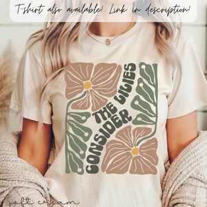 K&ouml;nnte beinhalten: Ein wei&szlig;es T-Shirt mit einem floralen Design und dem Text "CONSIDER THE LILIES" in einer Retro-Schrift.
