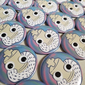 Pode incluir: Uma coleção de botões redondos e coloridos com uma ilustração em estilo cartoon. Cada botão apresenta um personagem bege claro estilizado com grandes olhos pretos e uma barba branca pontilhada, sobre um fundo azul, rosa e roxo.