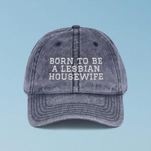 May include: A denim-colored baseball cap with the text "BORN TO BE A LESBIAN HOUSEWIFE" in white block letters. The cap has a curved brim and a button on top. The background is a light blue.
