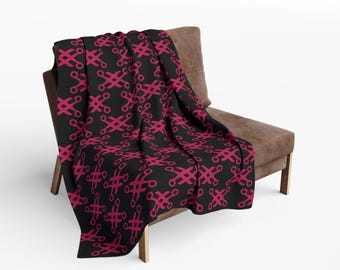 Op de afbeelding: Een zwarte deken met een herhalend patroon van magenta vormen, gedrapeerd over een bruine stoel. De vormen lijken op gestileerde scharen of kruisachtige ontwerpen, wat een gedurfd contrast creëert.