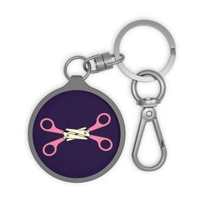May include: A round keychain with a dark purple background and a pink scissors design. The scissors are crossed and have a cream-colored handle. The keychain has a silver-colored clasp and key ring.