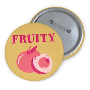 Może przedstawiać: Okrągła, żółta przypinka z napisem "FRUITY" w kolorze różowym. Poniżej ilustracja różowej brzoskwini z liściem i plasterkiem brzoskwini. Przypinka ma metalową agrafkę z tyłu.