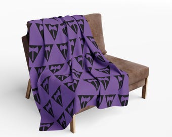 Op de afbeelding: Een paarse sprei met een herhaald patroon van zwarte bijlontwerpen, gedrapeerd over een bruine stoel. De sprei is gemaakt van een zacht materiaal, geschikt voor een woonkamer of slaapkamer. De stoel is bruin.