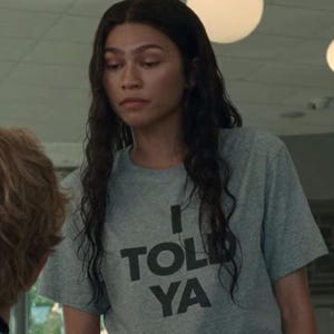 Könnte beinhalten: Ein graues T-Shirt mit dem Text "I TOLD YA" in schwarzen Blockbuchstaben. Das kurzärmelige Shirt scheint aus einem weichen Material zu sein. Die Person, die das Shirt trägt, hat lange, dunkle Haare.