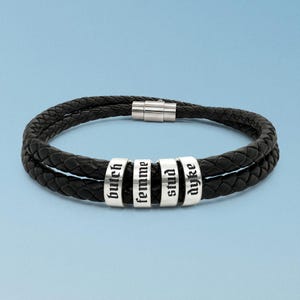 May include: A double-strand black braided leather bracelet featuring four silver-toned cylindrical metal beads. Each bead is engraved with black text in a gothic font, reading "butch," "femme," "stud," and "dyke." The bracelet includes a silver-toned magnetic clasp.