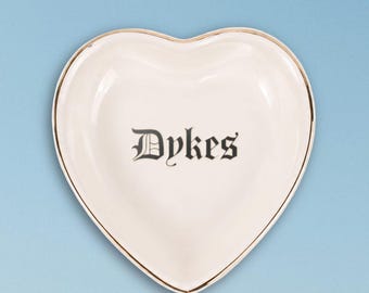 May include: A heart-shaped ceramic trinket dish in off-white with a metallic gold-toned rim. The center of the dish features the word Dykes printed in a black gothic-style font. The dish has a smooth, glossy finish and a curved edge.