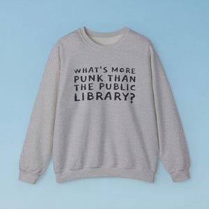 May include: A heather gray sweatshirt with the text "WHAT'S MORE PUNK THAN THE PUBLIC LIBRARY?" in black. The crew neck sweatshirt is a casual style, perfect for everyday wear. The background is a light blue.