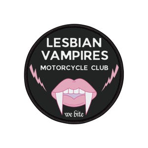 Peut inclure: Un écusson rond noir avec le texte blanc "LESBIAN VAMPIRES MOTORCYCLE CLUB". Il présente une bouche rose avec des crocs et les mots "we bite" en dessous. Des éclairs roses sont de chaque côté de la bouche.